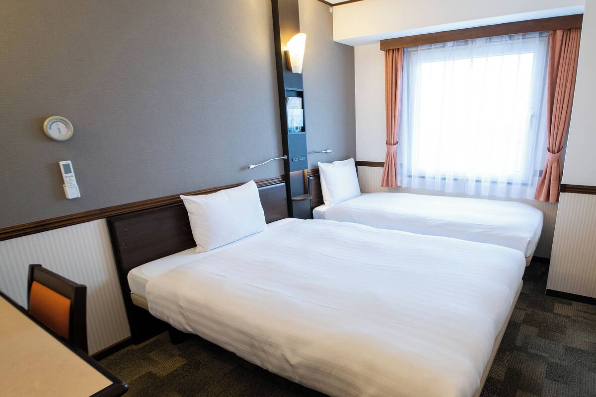 A hotel room with two beds, a nightstand, a lamp, a window with curtains, and a thermostat on the wall.