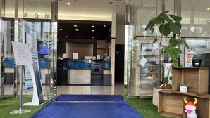 A hotel reception area with a blue carpet leading to the entrance, a glass door, and a reception desk with a computer and a plant. - Toyoko Inn Hikone Station Higashi (Hikone)