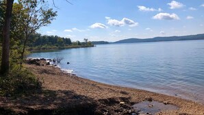 Ein ruhiger See mit einer felsigen Uferlinie, umgeben von Bäumen und Hügeln im Hintergrund.