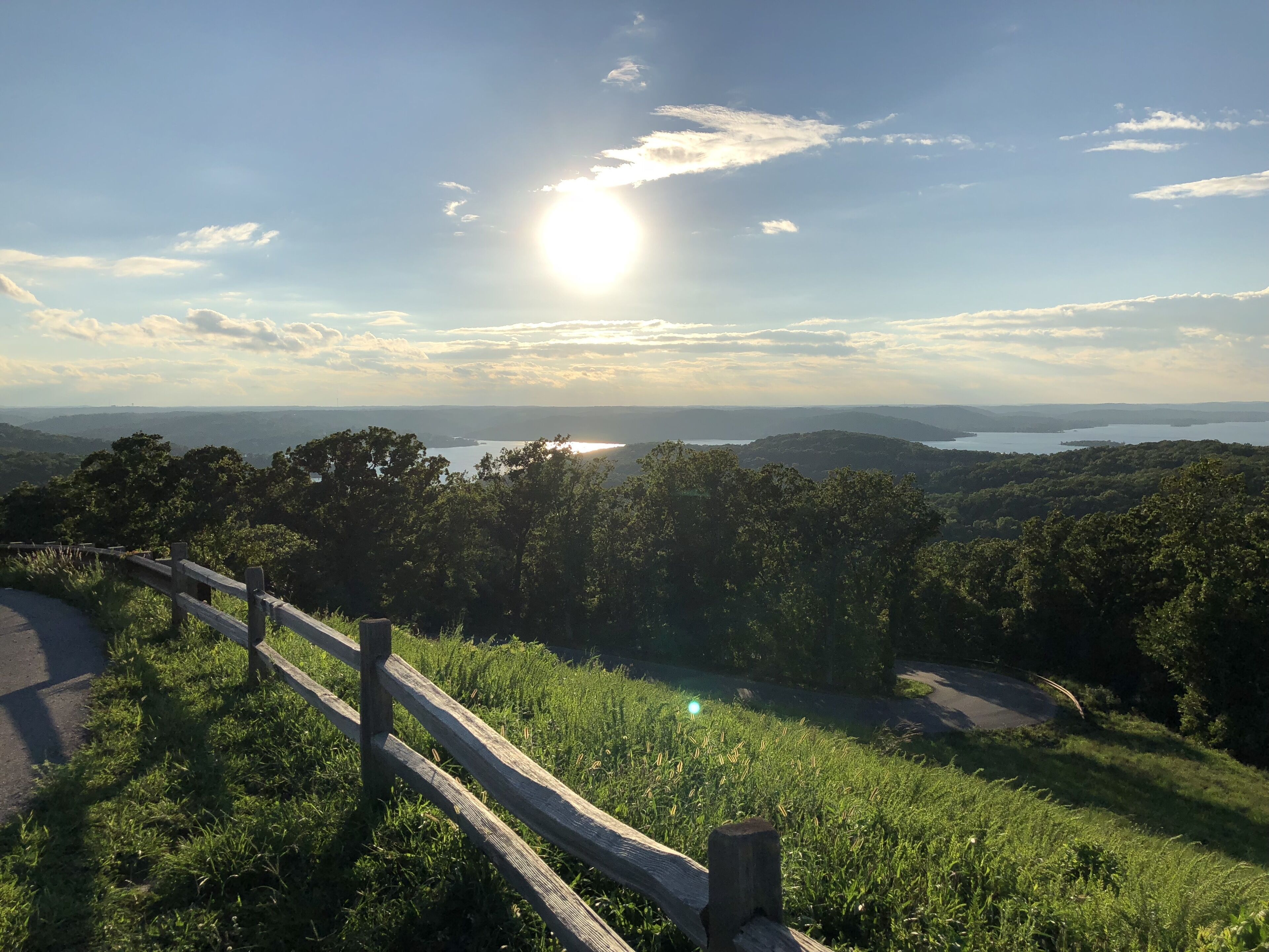 A scenic overlook with a wooden fence, lush greenery, and a distant view of a lake and mountains under a clear sky.