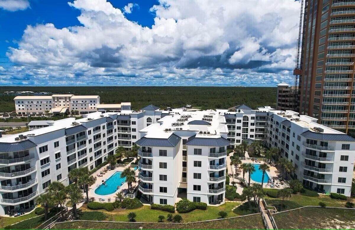 A multi-story hotel with a pool area, surrounded by greenery and a clear sky.