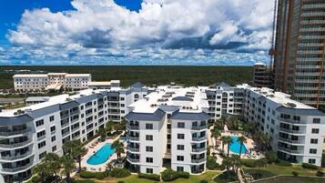 A multi-story hotel with a pool area, surrounded by greenery and a clear sky.