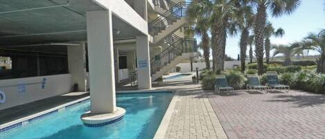 A swimming pool with a brick walkway, palm trees, and a multi-story building with balconies.