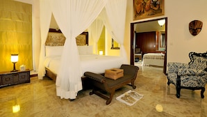 A bedroom with a four-poster bed, a wooden bench, a patterned armchair, and a nightstand with a lamp. - Puri Mas Spa Resort (Senggigi)