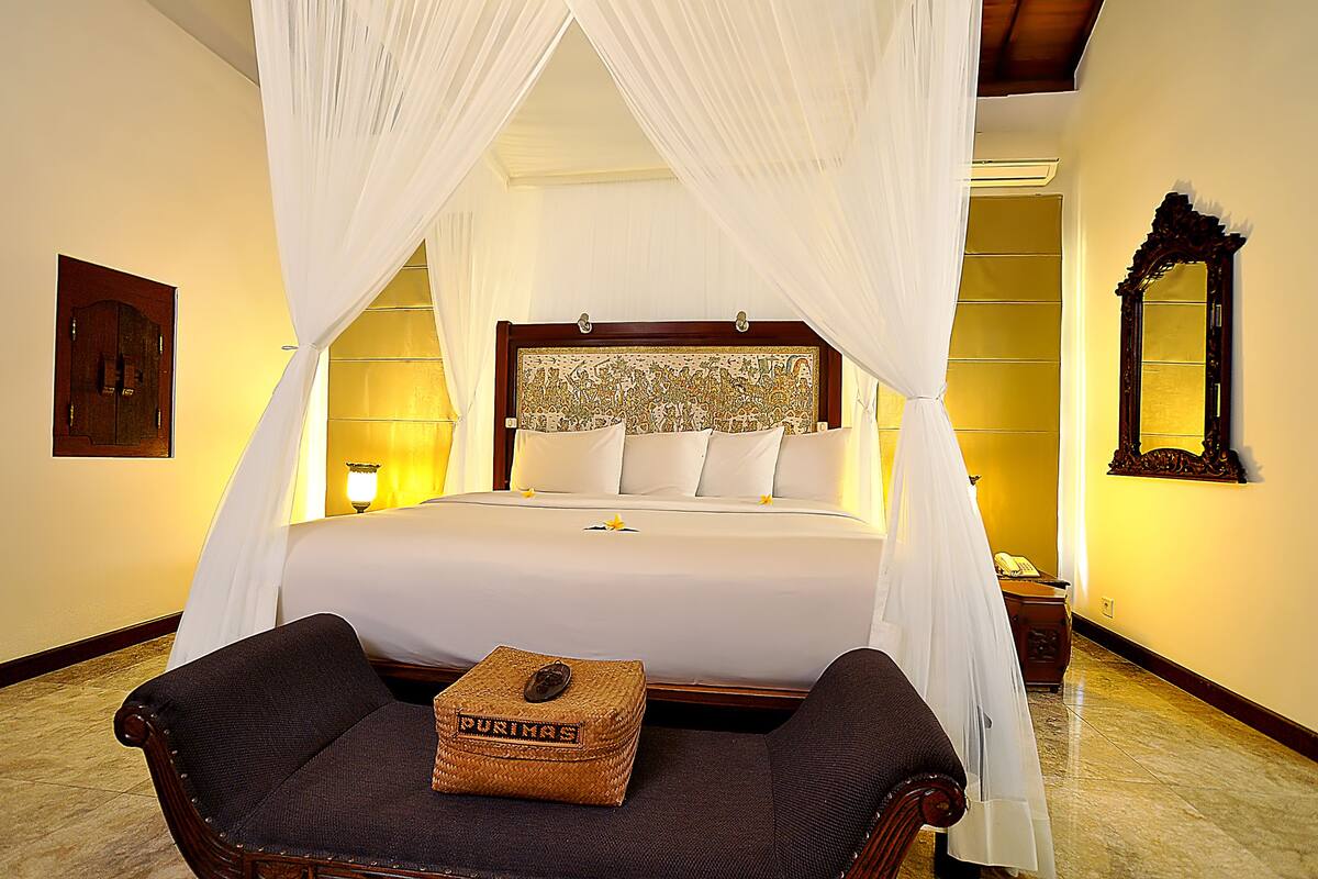 A hotel room with a four-poster bed, a nightstand, a lamp, and a chair with a woven basket.