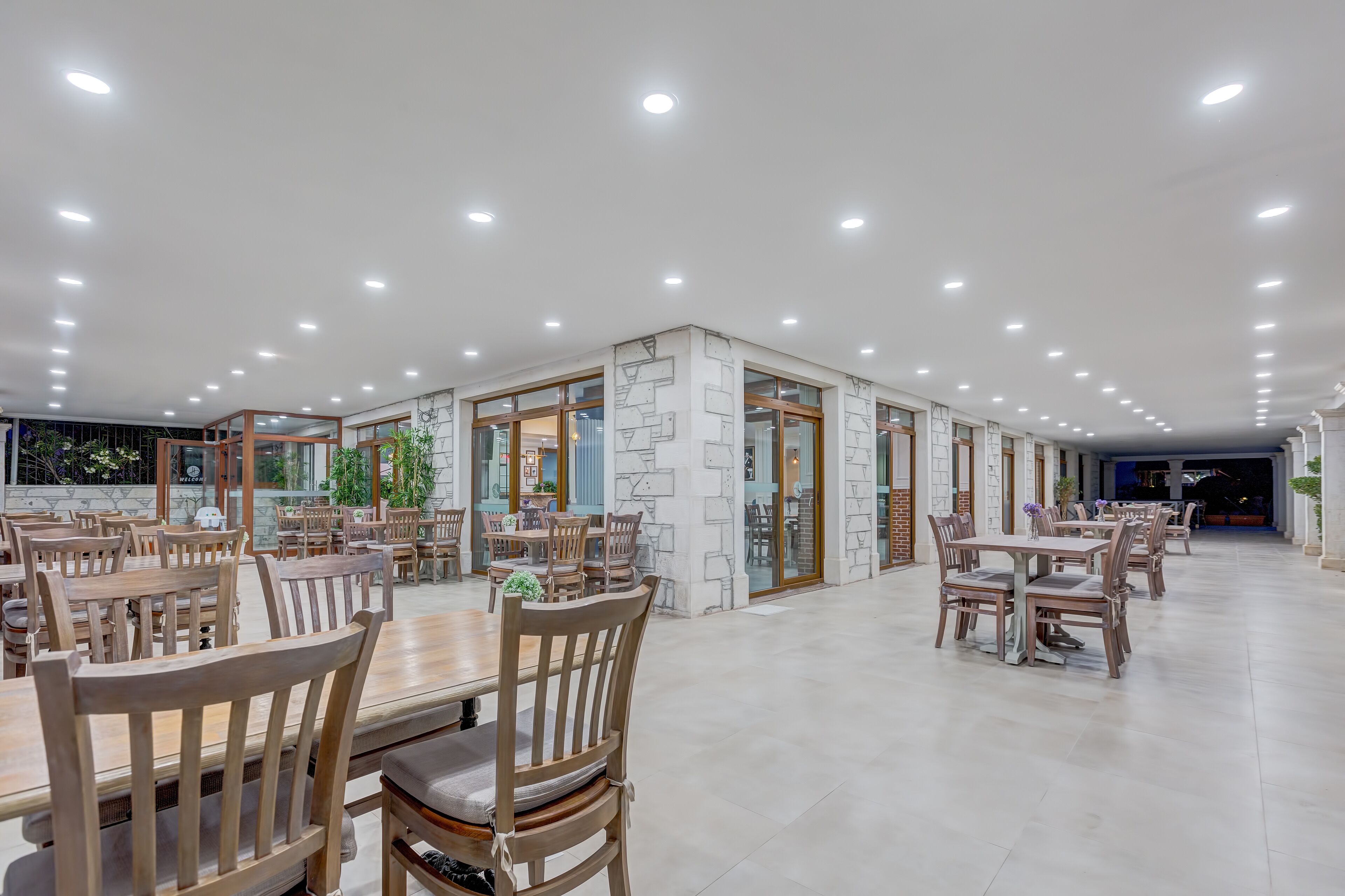 Un comedor amplio con varias mesas y sillas de madera, luces empotradas en el techo y grandes ventanales que dejan entrar luz natural.