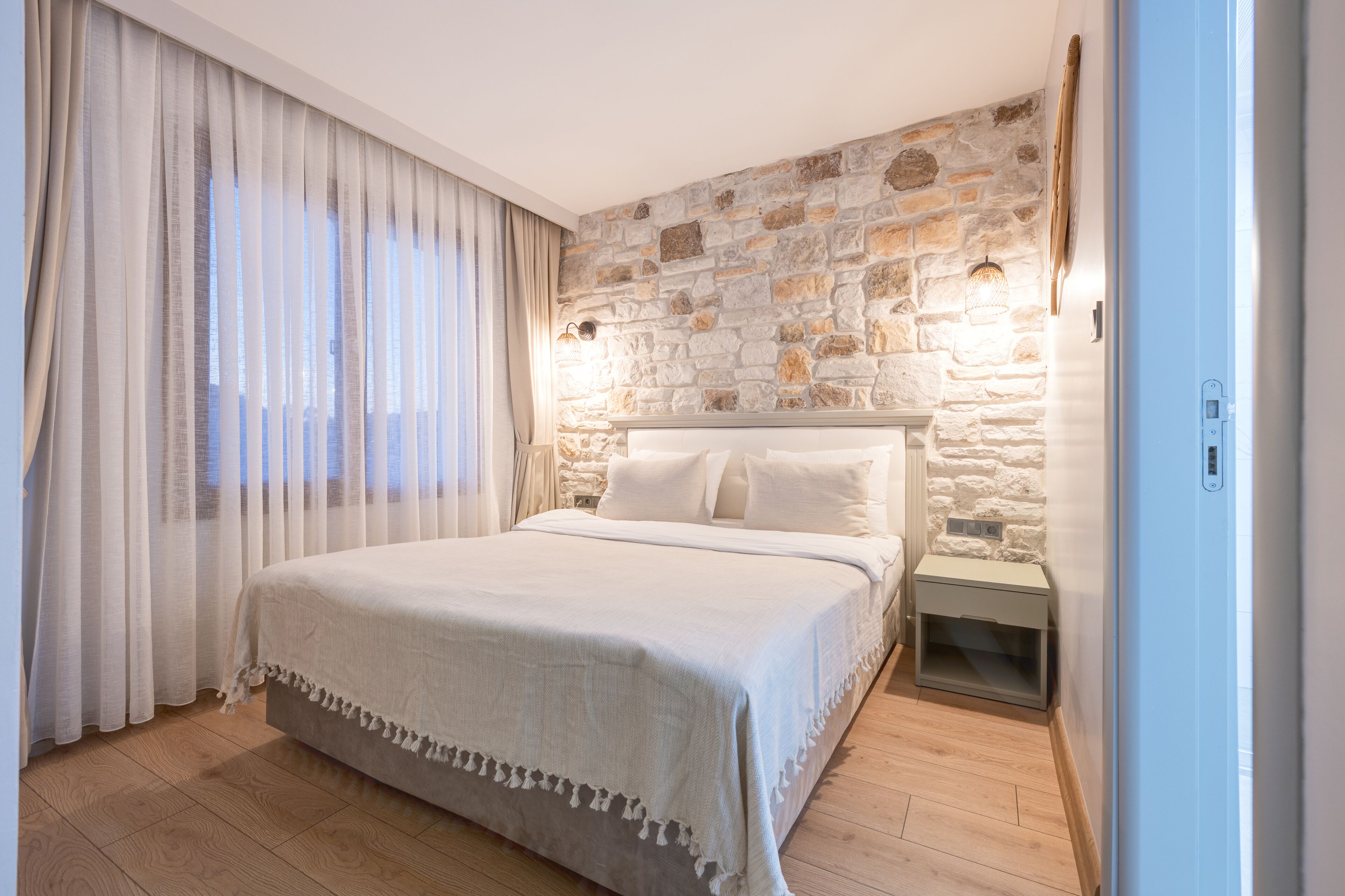 A bedroom with a stone wall, a bed with white linens, a nightstand, and a window with sheer curtains.