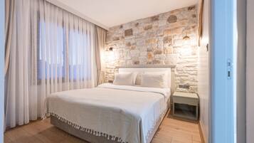 A bedroom with a stone wall, a bed with white linens, a nightstand, and a window with sheer curtains.