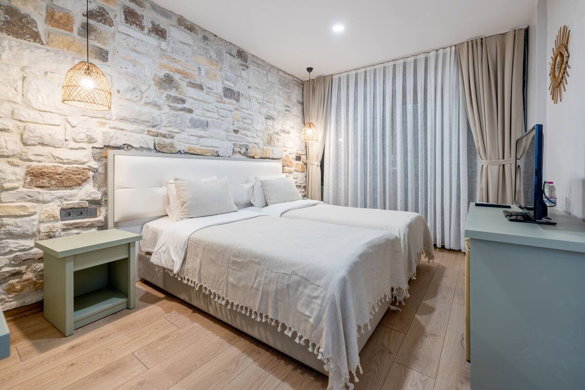 Ein Schlafzimmer mit Steinwand, einem Bett mit weißer Bettwäsche, einem Nachttisch, einer Lampe und einem Fernseher auf einem Standfuß.