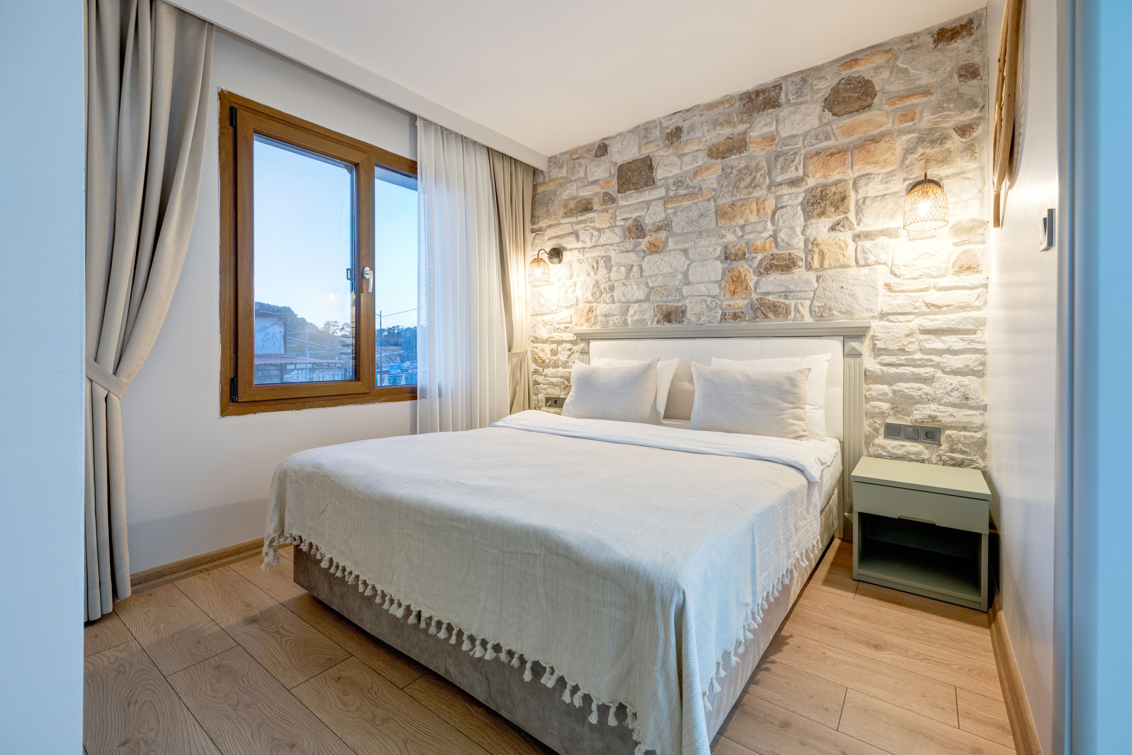 A bedroom with a stone wall, a large bed, a nightstand, and a window with curtains.