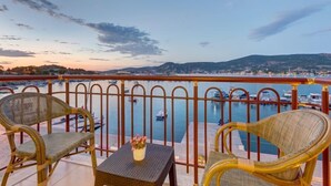 Un balcón con sillones de mimbre y una mesa pequeña, con vistas a un puerto deportivo con barcos y un paisaje urbano al fondo.