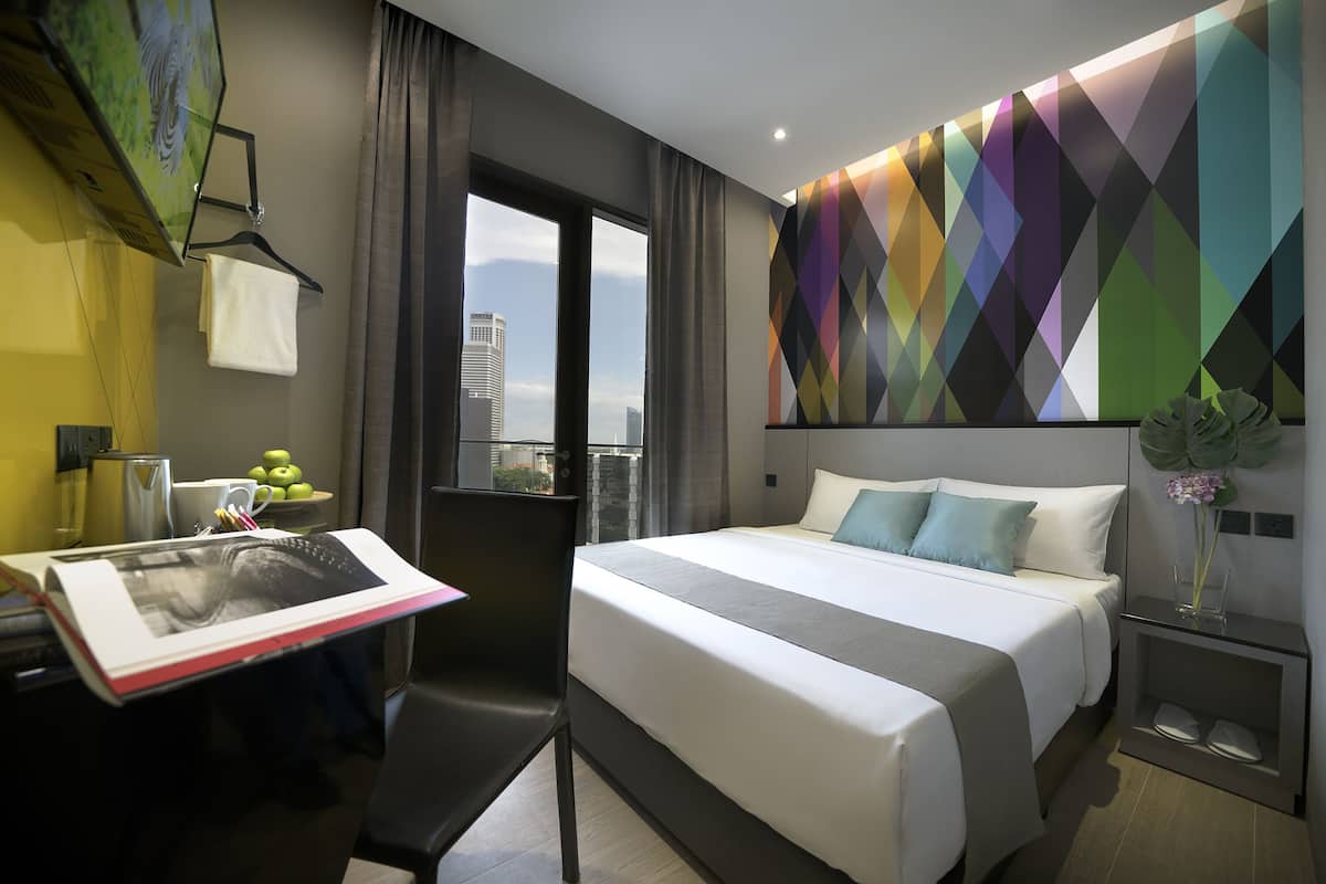 A modern hotel room with a large bed, a desk with a flat-screen TV, a city view, and a colorful geometric wall design.