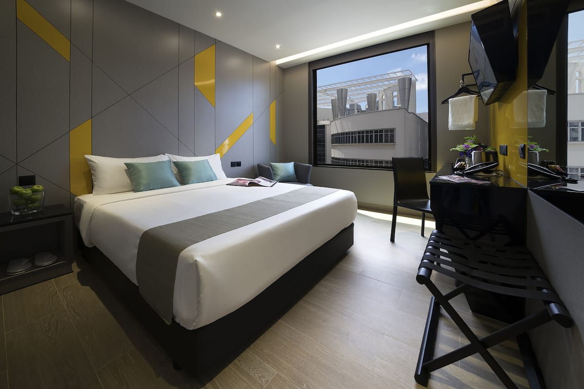 A modern hotel room with a large bed, a desk, and a view of the cityscape through a large window.
