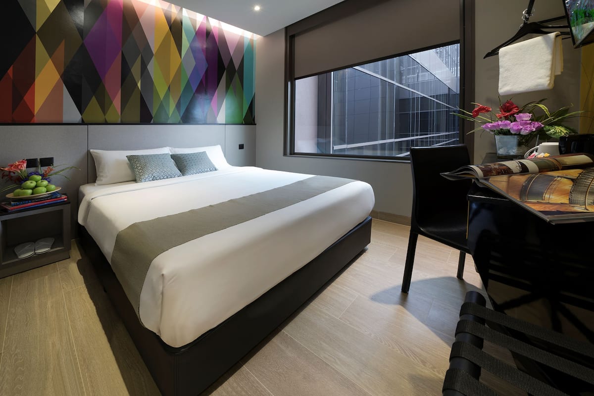 A modern hotel room with a large bed, a desk with a chair, a window with a view of the city, and a colorful wall design.