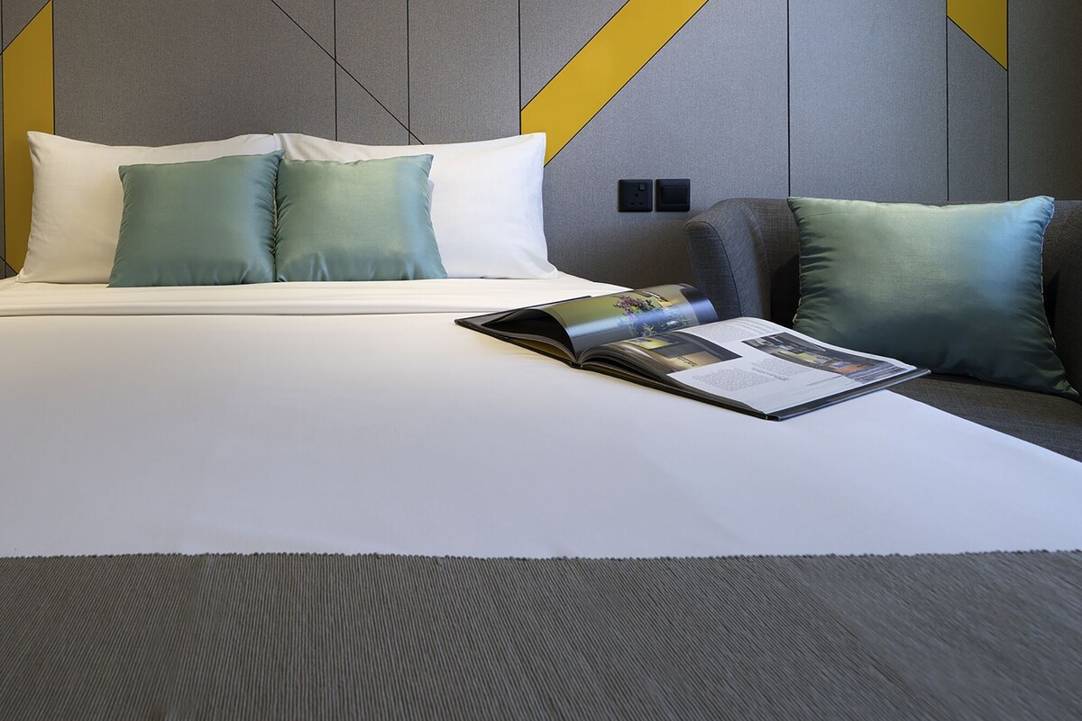 Una cama bien hecha con almohadas blancas y color turquesa, una revista sobre la cama y un diseño de habitación moderno con patrones geométricos en la pared.
