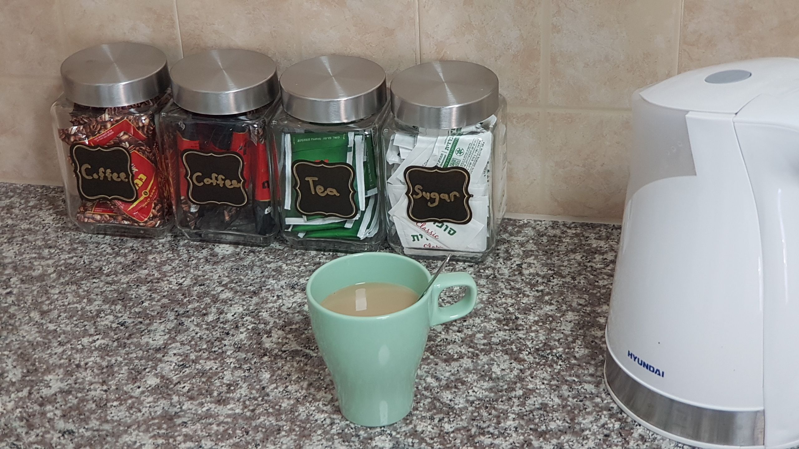 Cuatro frascos de vidrio con tapas de metal etiquetados como 'Coffee', 'Tea' y 'Sugar' sobre una encimera de cocina, una taza verde con una cuchara y una cafetera blanca.