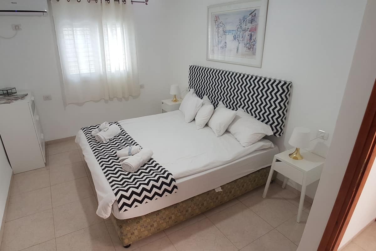 A neatly arranged bedroom with a bed, bedside tables, a small desk, and a wall-mounted air conditioner.