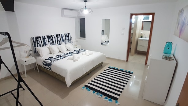 A bedroom with a bed, a bedside table, a nightstand, a hanging light fixture, a striped rug, a mirror, and a door leading to a bathroom. - Hili Apartments (Tiberias)