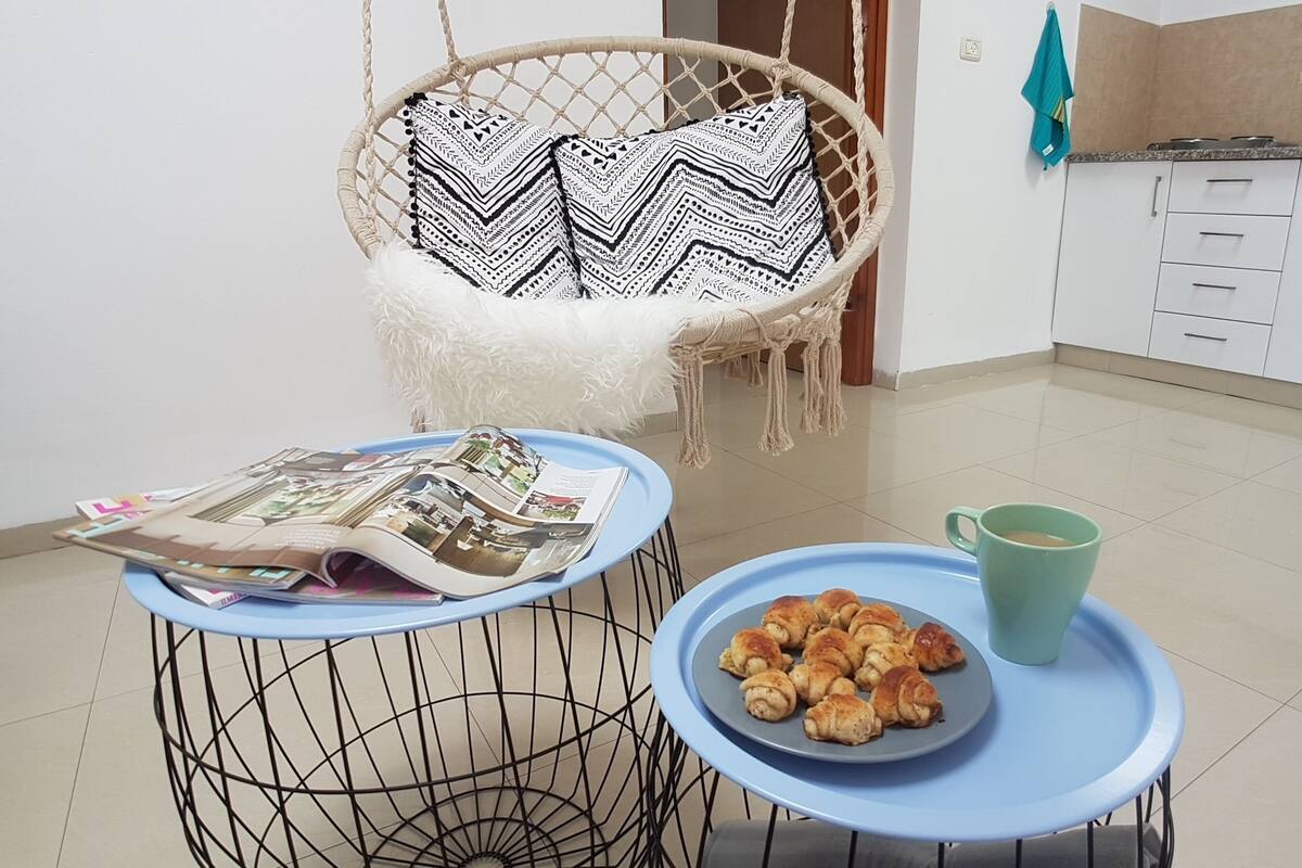A kitchen with a hanging chair, a table with a magazine, a plate of pastries, and a mug.