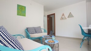 A modern living room with a sofa, a small round table with a laptop and pastries, and a dining area with chairs. - Hili Apartments (Tiberias)
