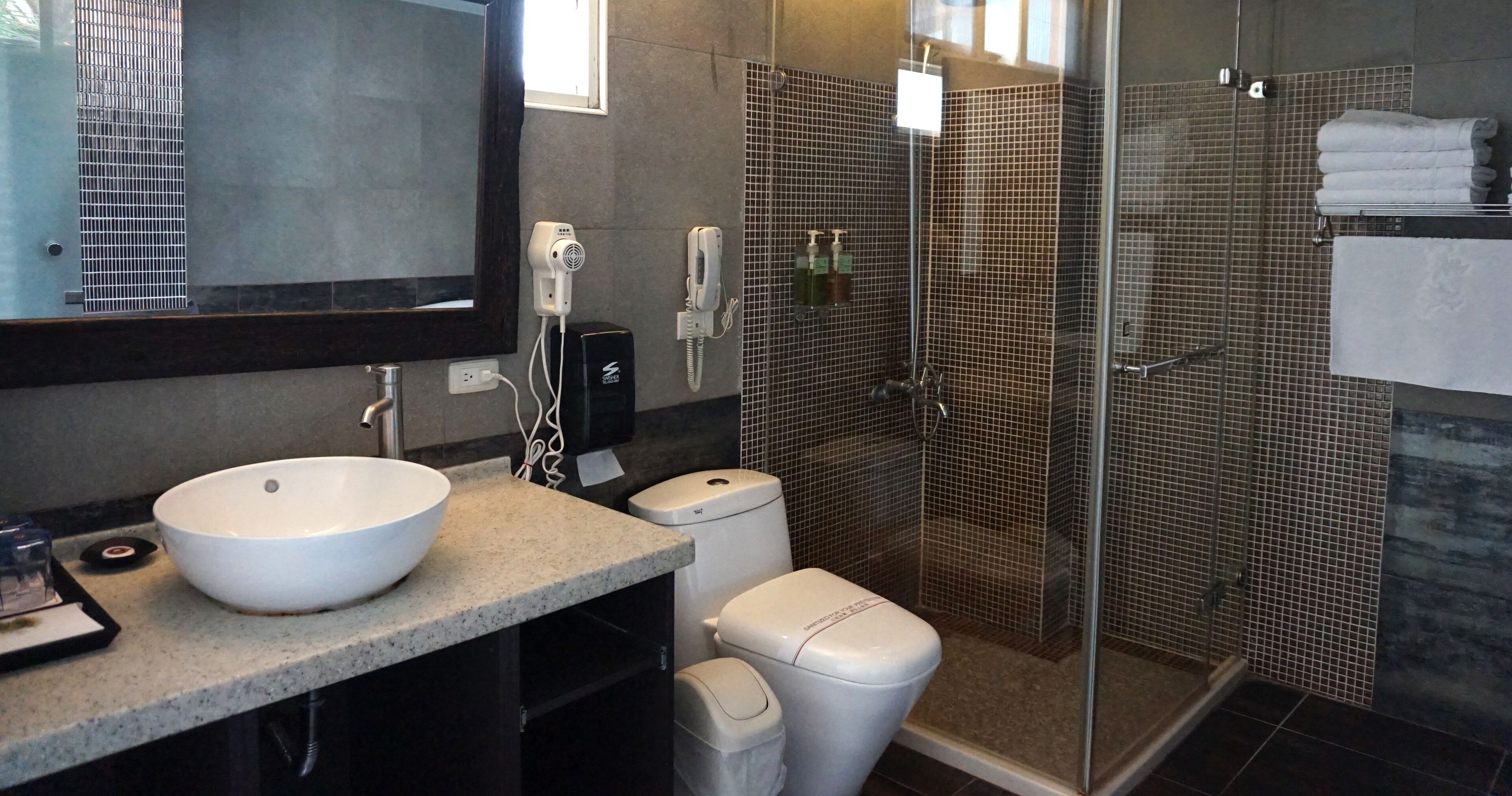 Un baño con un lavamanos grande de color blanco, un inodoro, una ducha con mampara de vidrio y un espejo sobre el lavamanos.