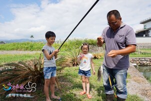 Un hombre y dos niños de pie al aire libre cerca de un cuerpo de agua, posiblemente un río o estanque, con follaje y un cielo despejado al fondo.