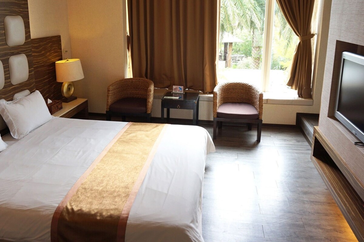 Habitación de hotel con una cama grande, dos sillones de mimbre, un televisor y una ventana con cortinas.