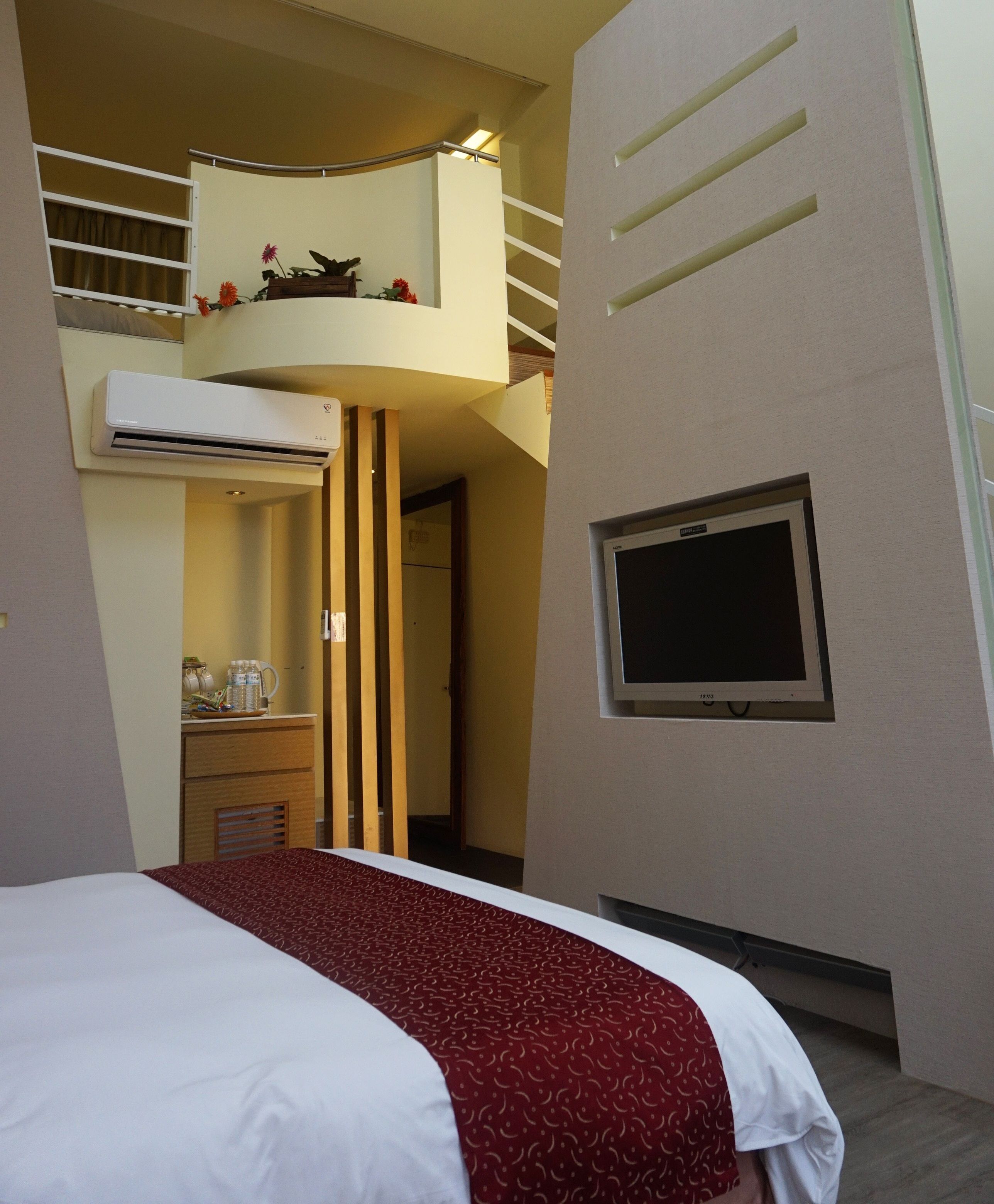 A hotel room with a bed, a TV mounted on the wall, a balcony with plants, and an air conditioning unit.