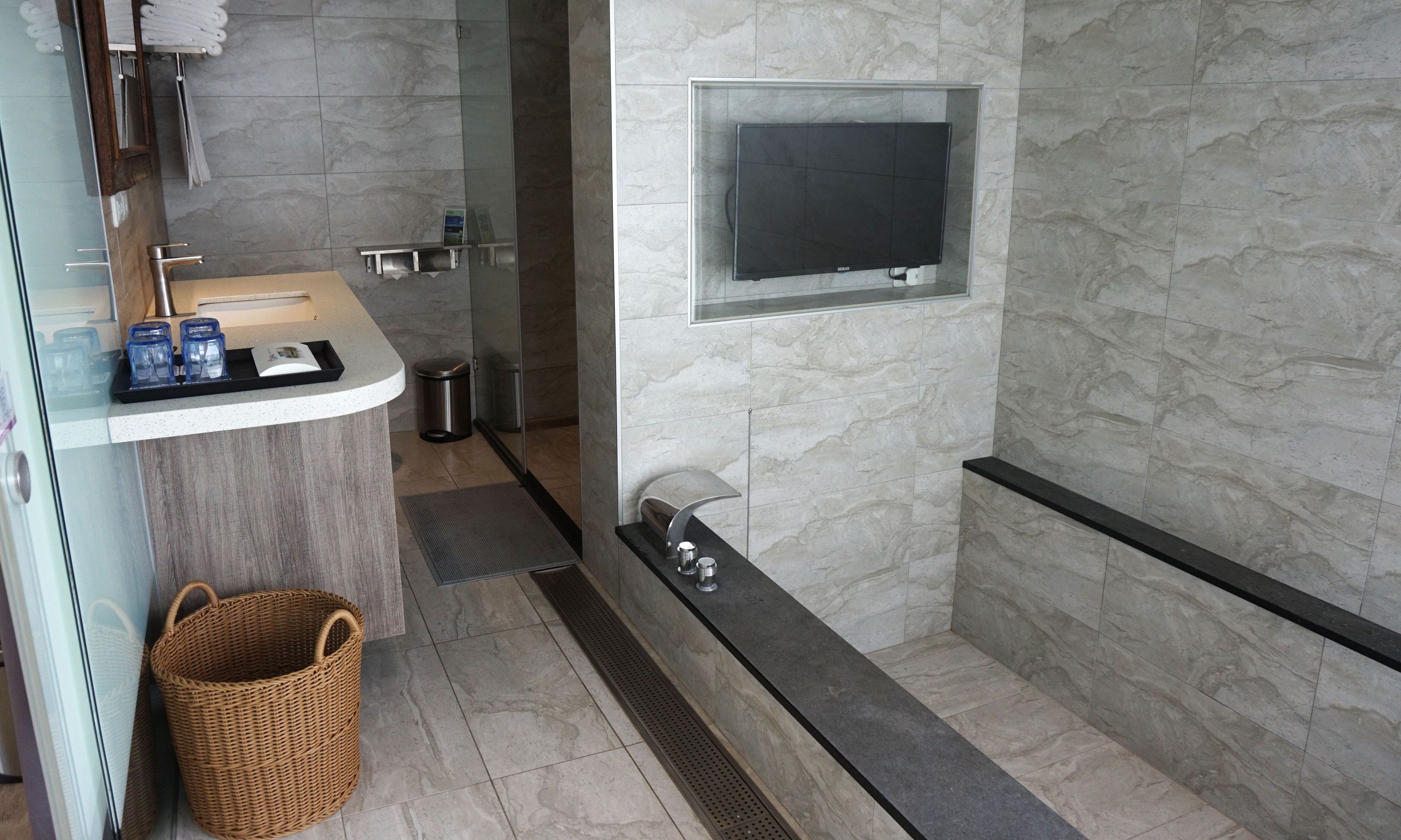 Un baño moderno con pared de mármol, cabina de ducha de vidrio, televisor montado y una cesta de mimbre.
