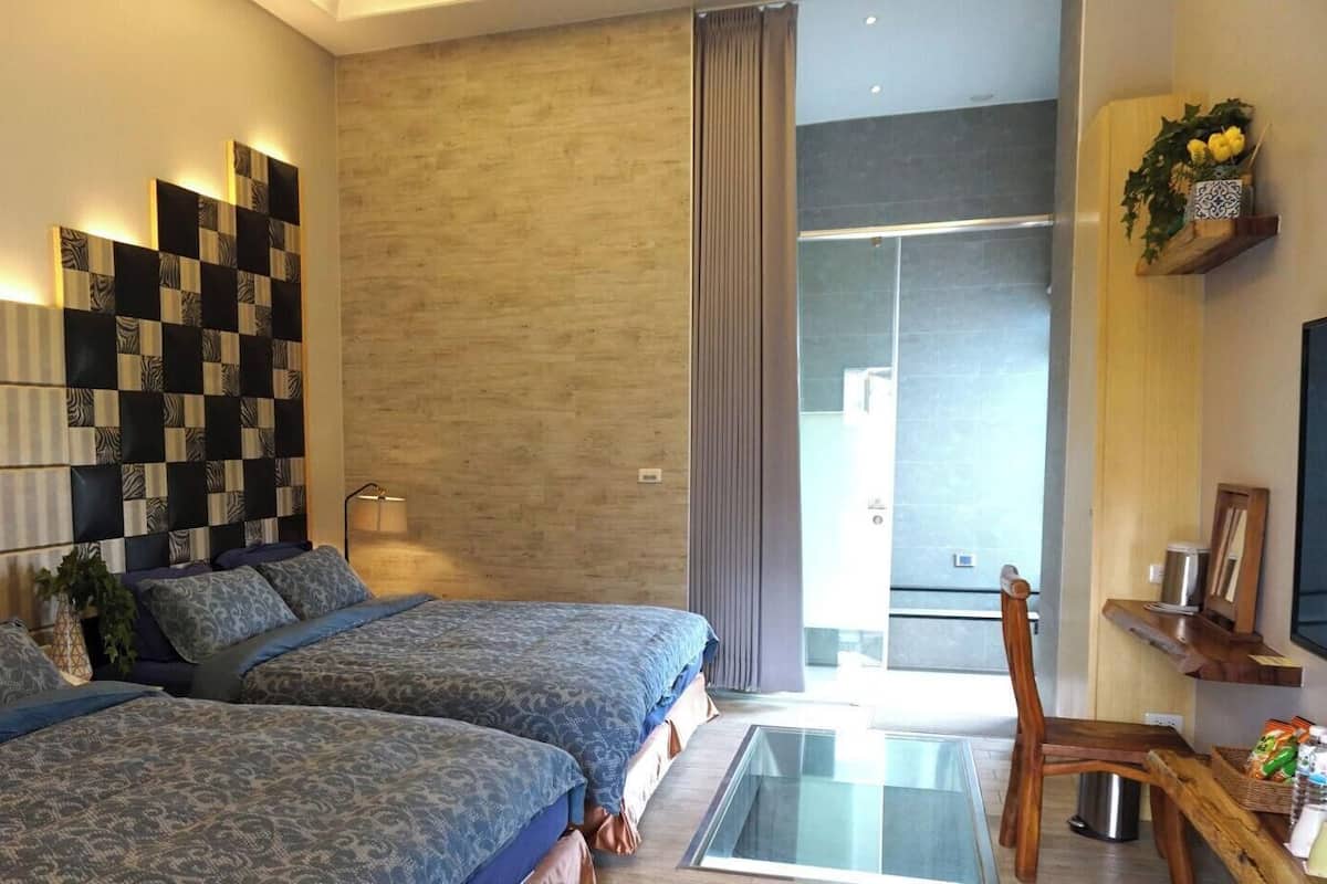 A modern bedroom with two beds, a wooden headboard, a glass table, and a view of the outdoors.