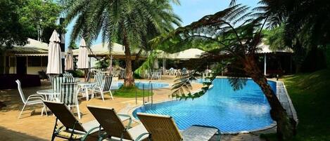 Zona junto a la piscina con sillones y sombrillas, rodeada de palmeras.