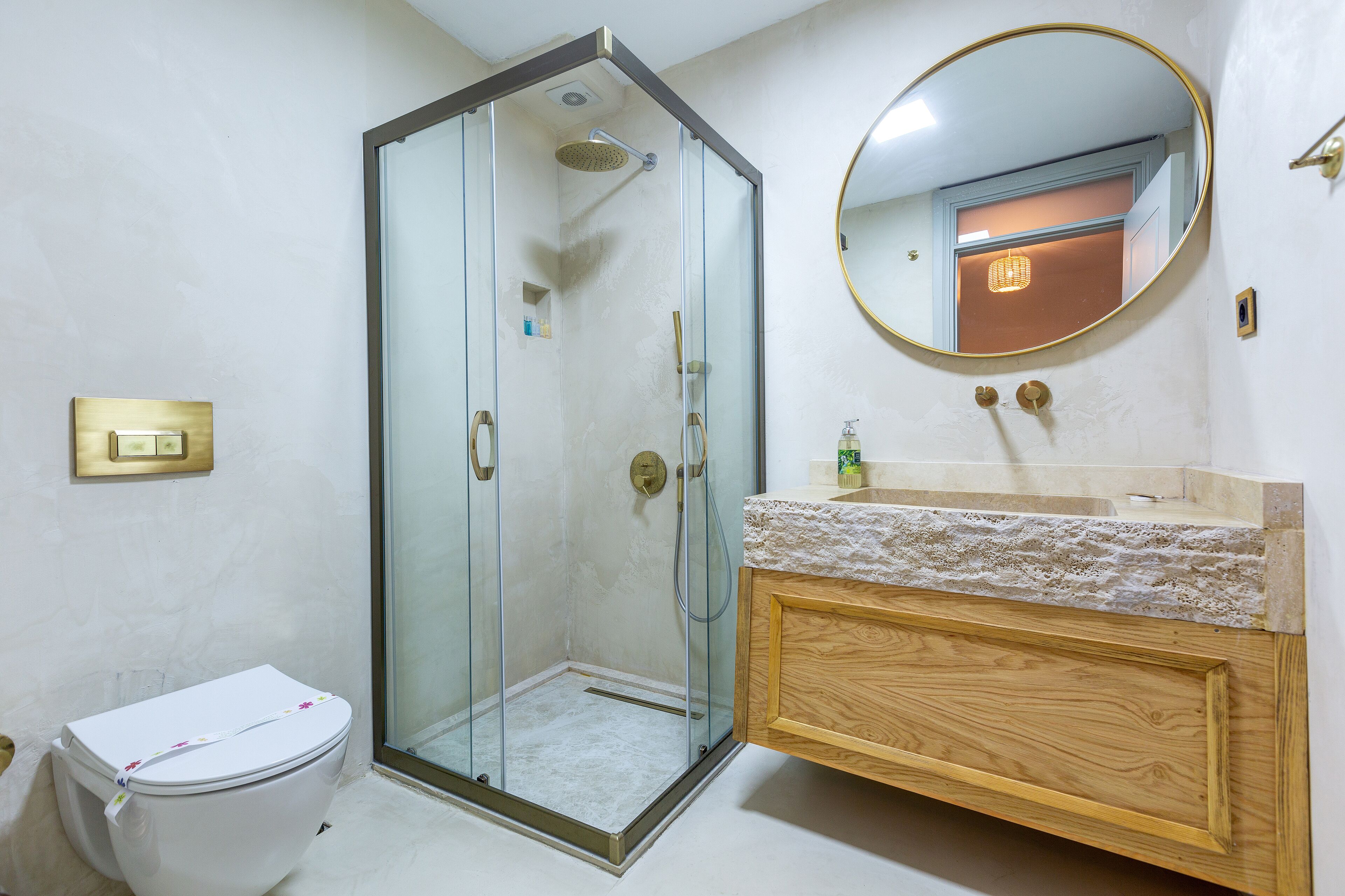 A bathroom with a glass-enclosed shower, a toilet, a wooden vanity with a marble countertop, and a round mirror.