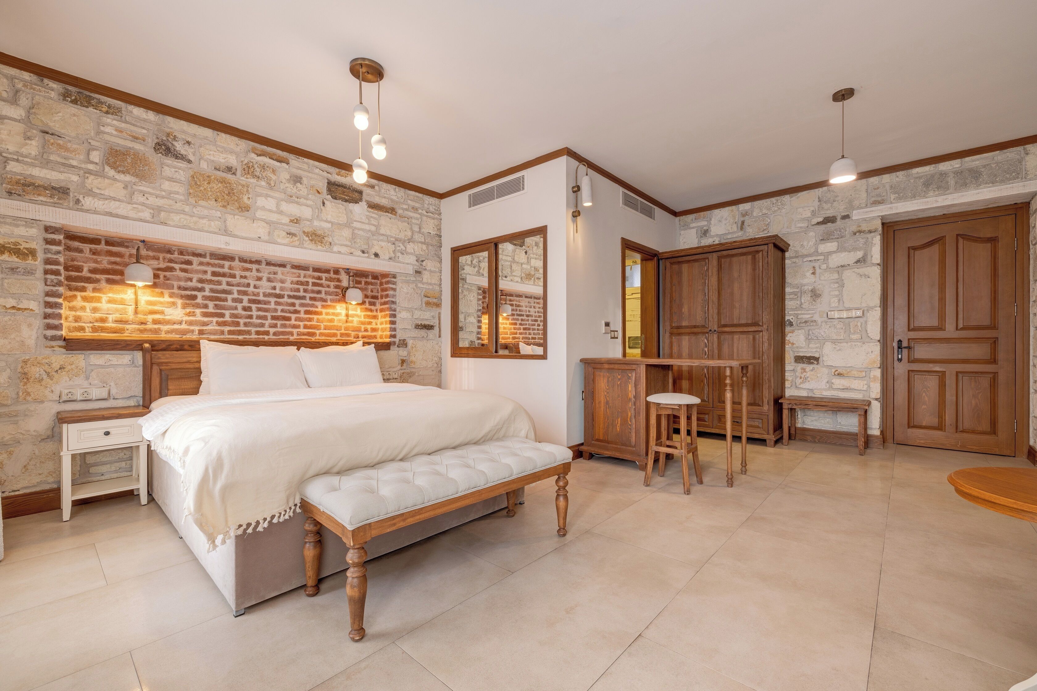 A bedroom with a large bed, a wooden bench, a nightstand, a wooden wardrobe, and a desk with a chair.