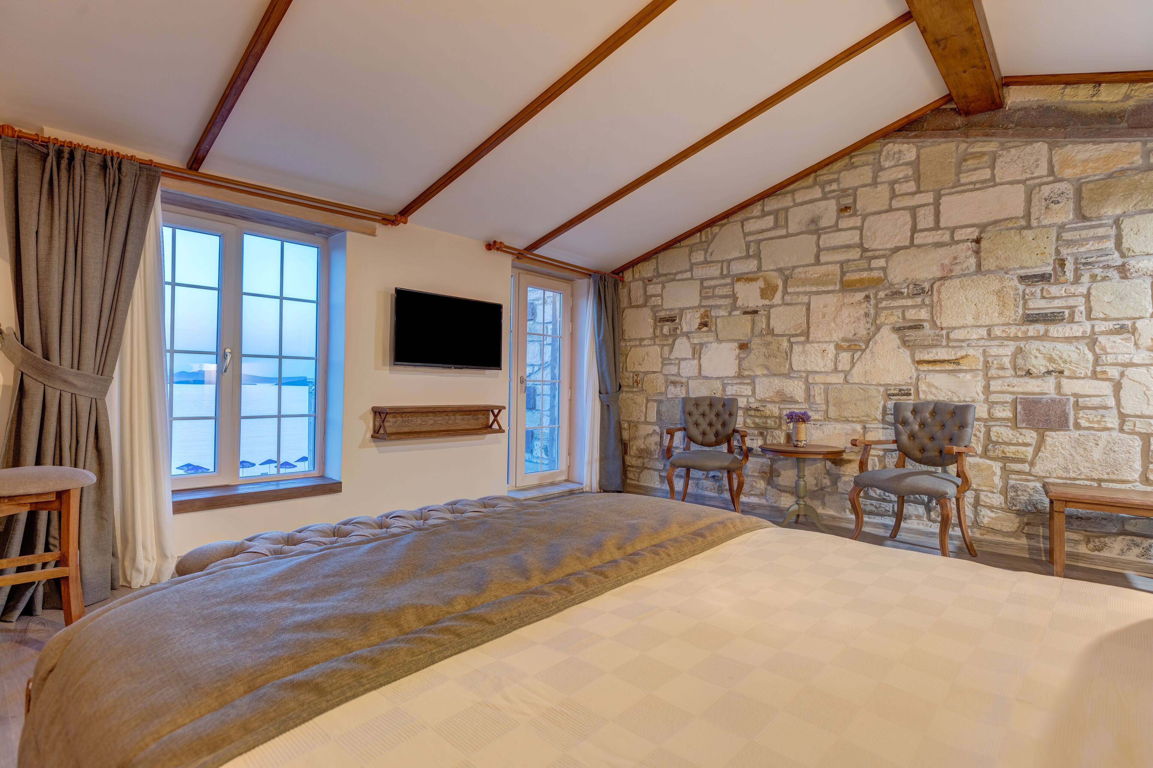 A bedroom with a stone wall, a bed, a television, and two chairs.
