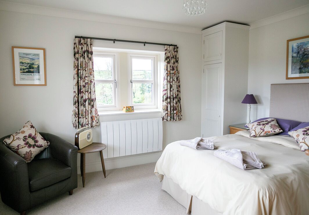 Une chambre à coucher avec un lit, une chaise, un radiateur, une fenêtre avec des rideaux, une lampe et un tableau encadré au mur.