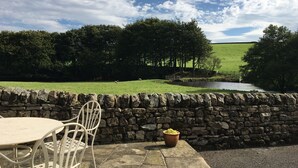 Eine Steinsäule mit einem Tisch, Stühlen, einem Blumentopf und Blick auf ein grünes Feld mit Bäumen und einem kleinen Gewässer.