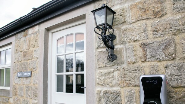 Ein Steinhaus mit einer weißen Tür, einem Schild mit der Aufschrift "Yates Cottage" und einer Ladestation für Elektrofahrzeuge, beschriftet mit "PROJECT EV".