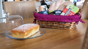 Ein Esstisch mit Kuchen auf einer Glasschale, ein Korbwesen mit Lebensmitteln und eine Küche im Hintergrund.