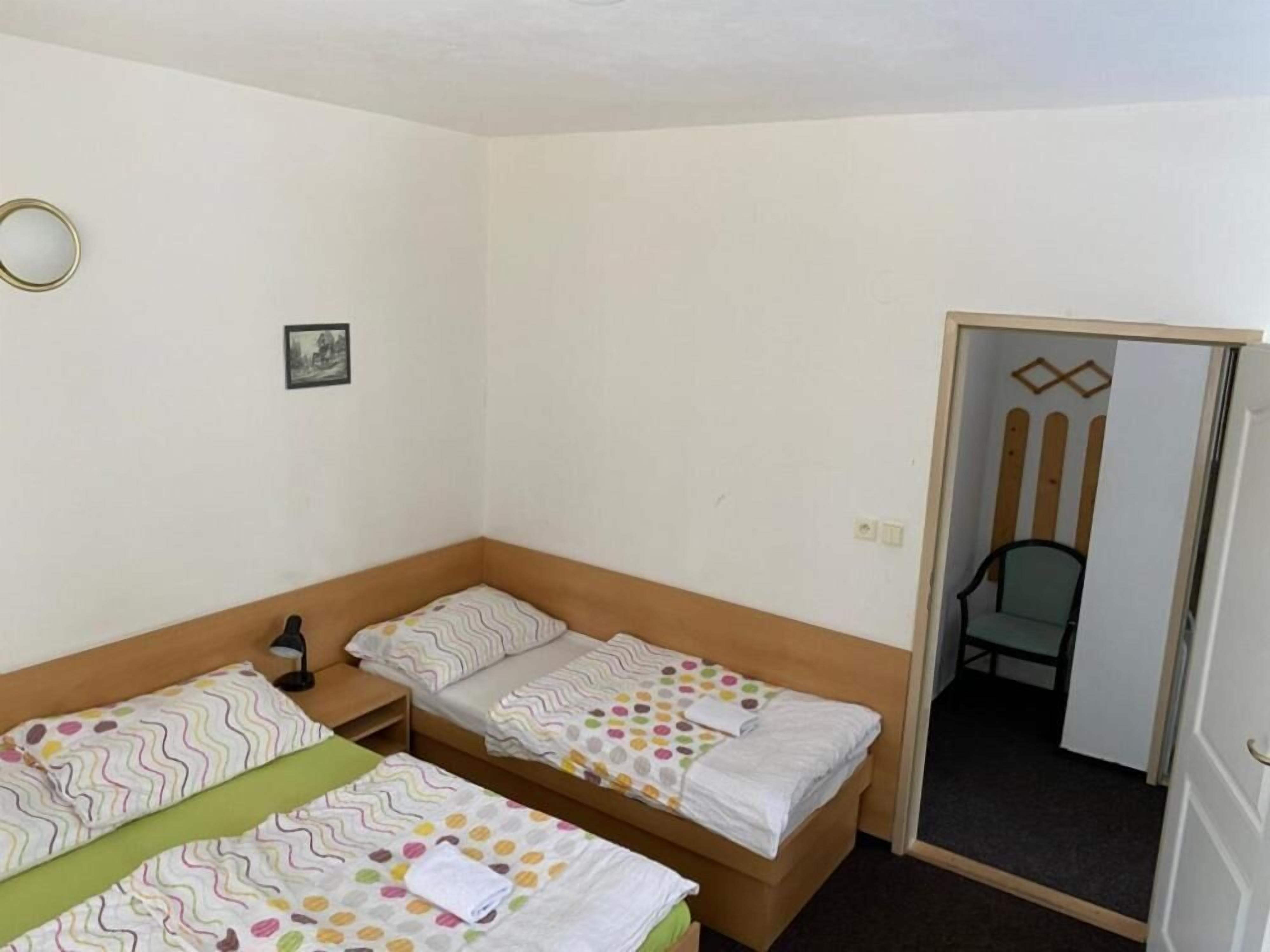 Una habitación pequeña y ordenada con dos camas individuales, una mesita de noche, una silla y una puerta que da a otra habitación.