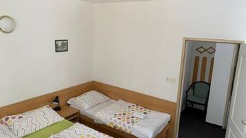 Una habitación pequeña y ordenada con dos camas individuales, una mesita de noche, una silla y una puerta que da a otra habitación.