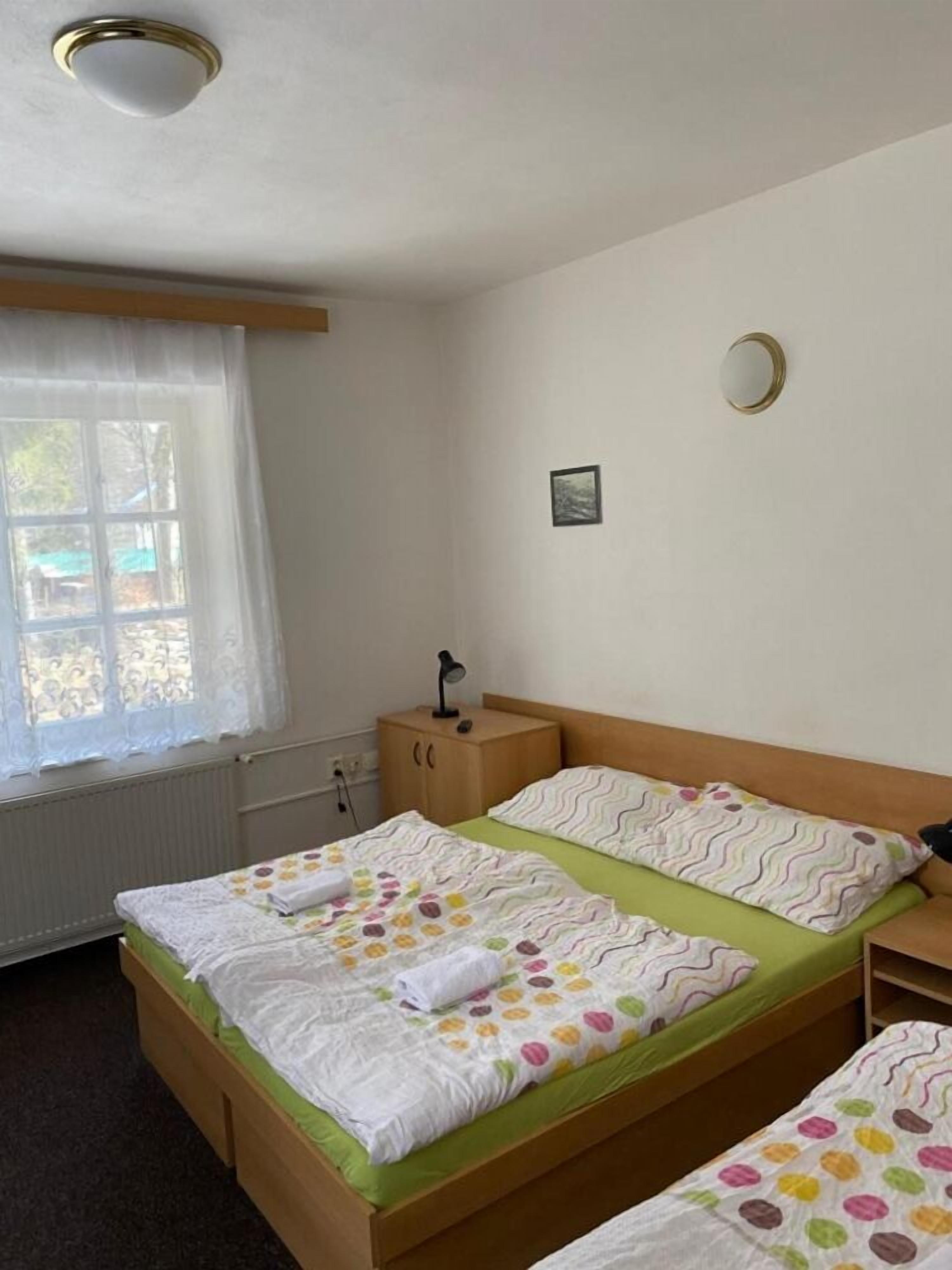 Ein Schlafzimmer mit zwei Betten, einem Fenster mit durchscheinenden Vorhängen, einem Nachttisch mit Lampe und einem kleinen Bild an der Wand.