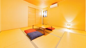 Une pièce traditionnelle avec un sol recouvert de tatamis, une petite table et des coussins, un appareil de climatisation et une petite fenêtre.