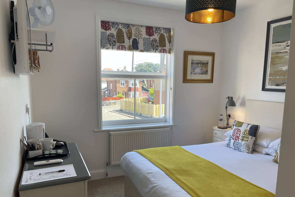 A bedroom with a bed, a desk with a computer, a window with a view of a residential area, and a bedside table with a lamp.