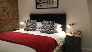 A neatly made bed with a red blanket, two patterned pillows, a black headboard, a framed picture on the wall, and bedside tables with lamps and water bottles. - Holway House (Sheringham)