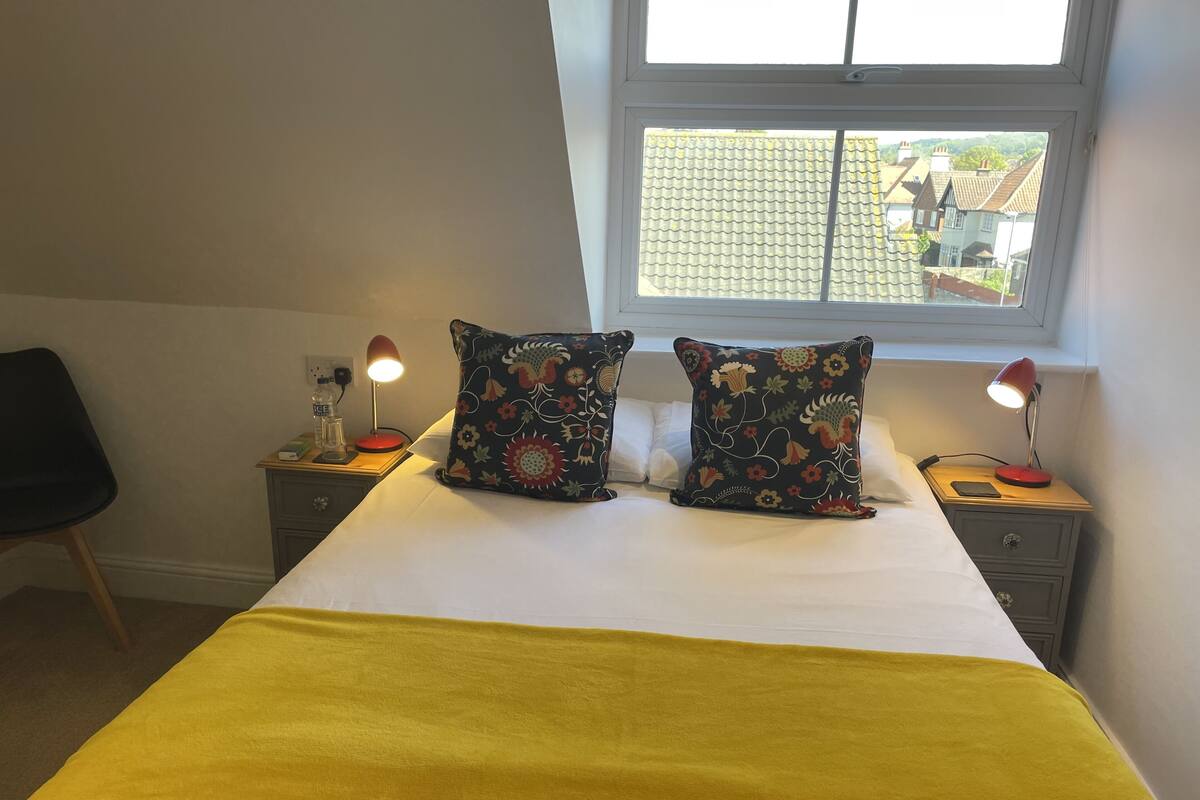 A neatly made bed with two decorative pillows, a bedside table with a lamp, and a window with a view of buildings.