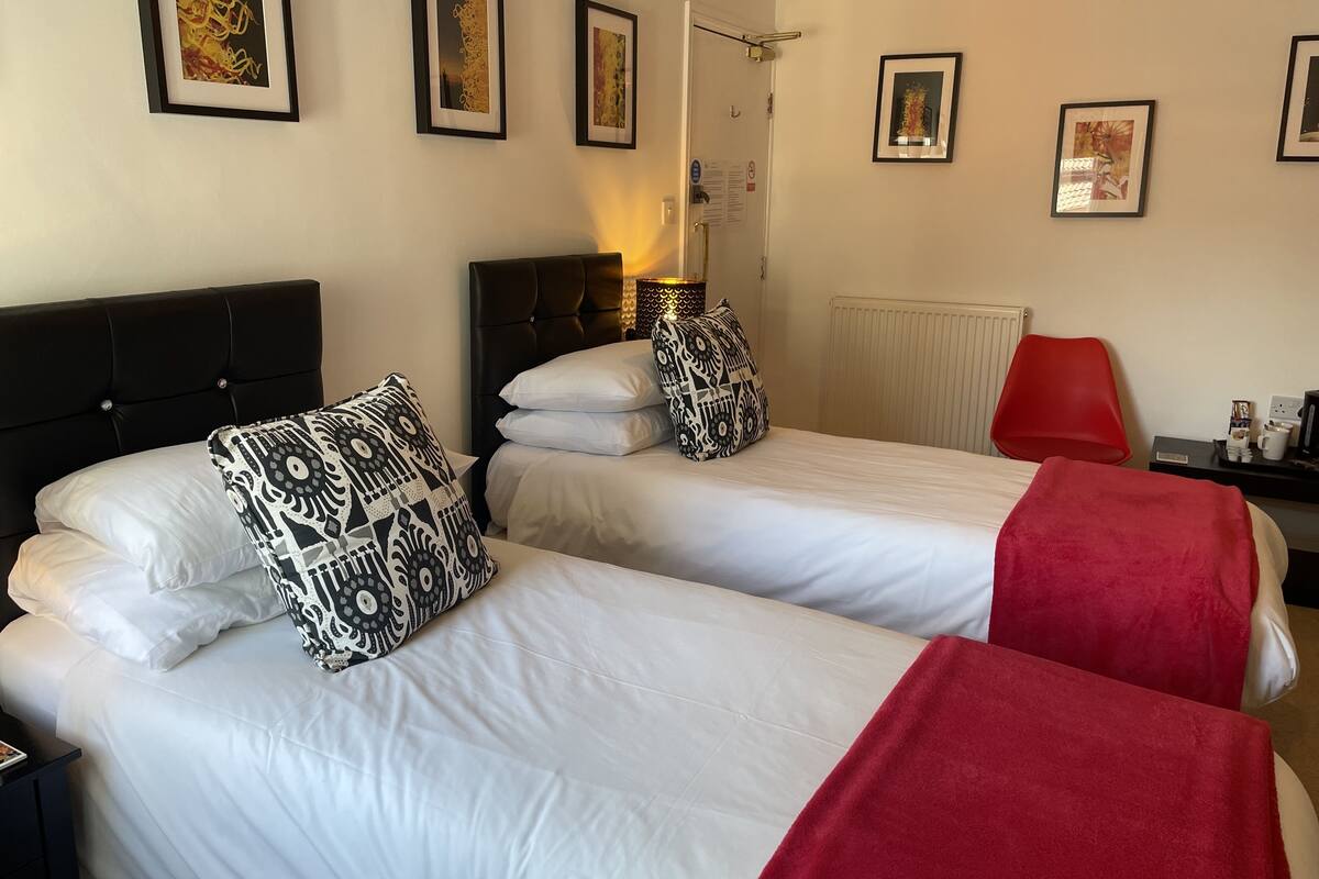 A hotel room with two beds, white bedding, patterned pillows, a red throw blanket, and framed artwork on the walls.