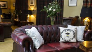 A plush, dark red leather sofa with white and black patterned cushions, positioned in a warmly lit room with plaid curtains, a tall vase with greenery, and framed artwork on the walls. - Glenegedale House (Islay Island)