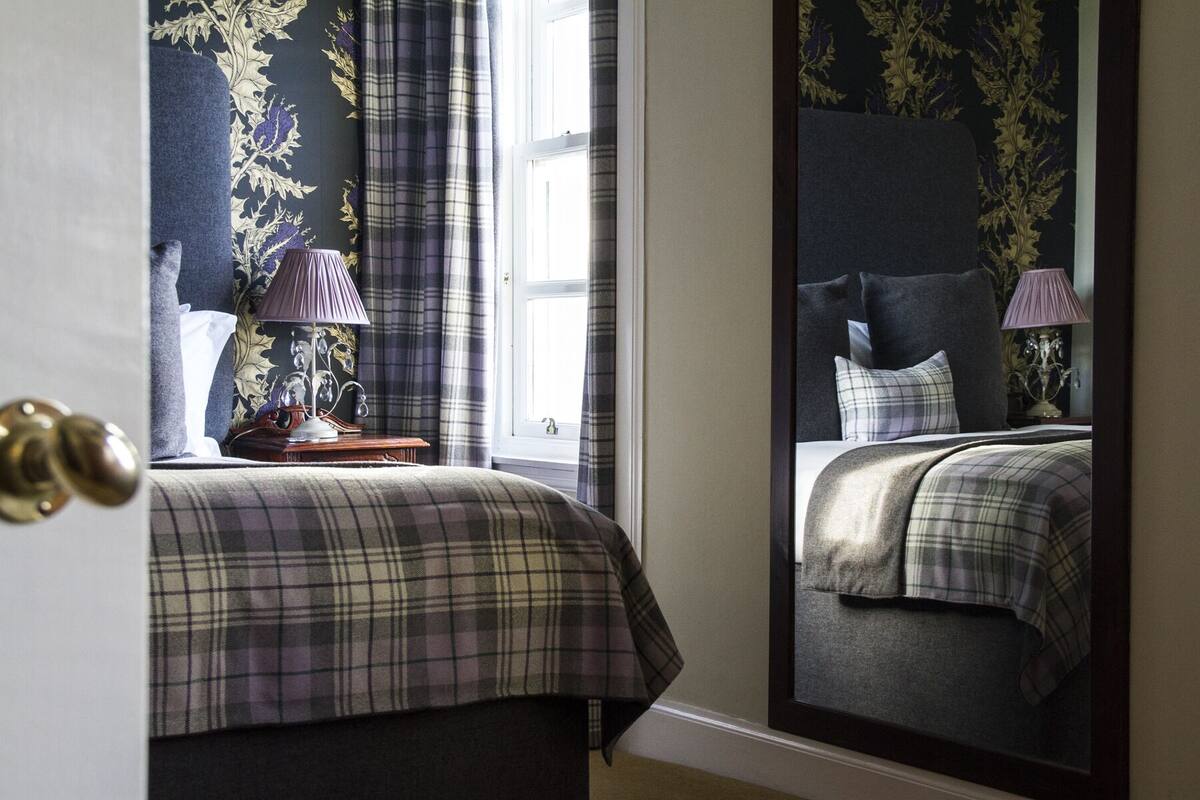 A bedroom with a large mirror, a bed with a plaid bedspread, a nightstand with a lamp, and a window with curtains.