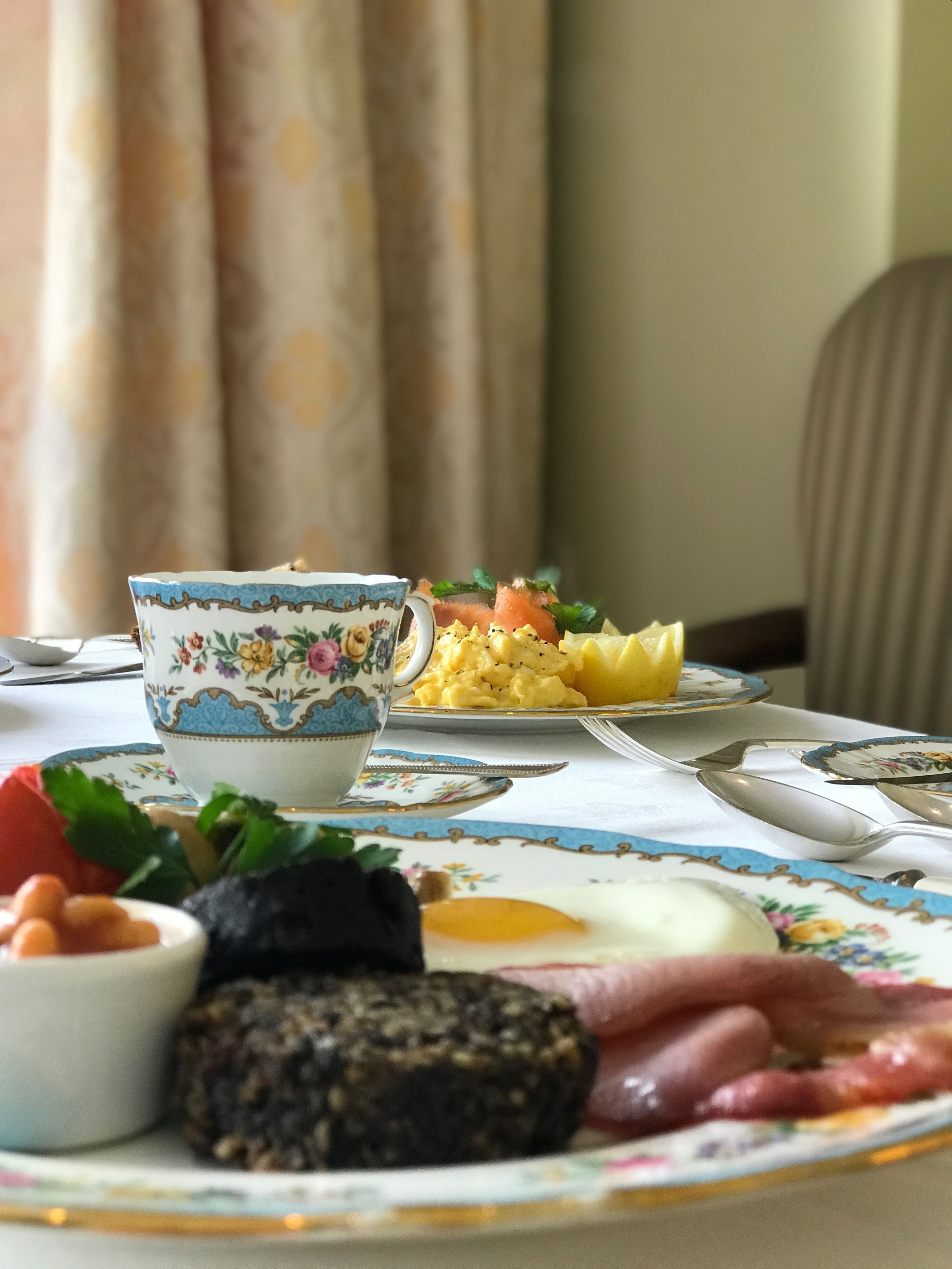 A table with a floral tablecloth, a bowl of soup, a plate of scrambled eggs, a slice of toast, and a piece of meat.