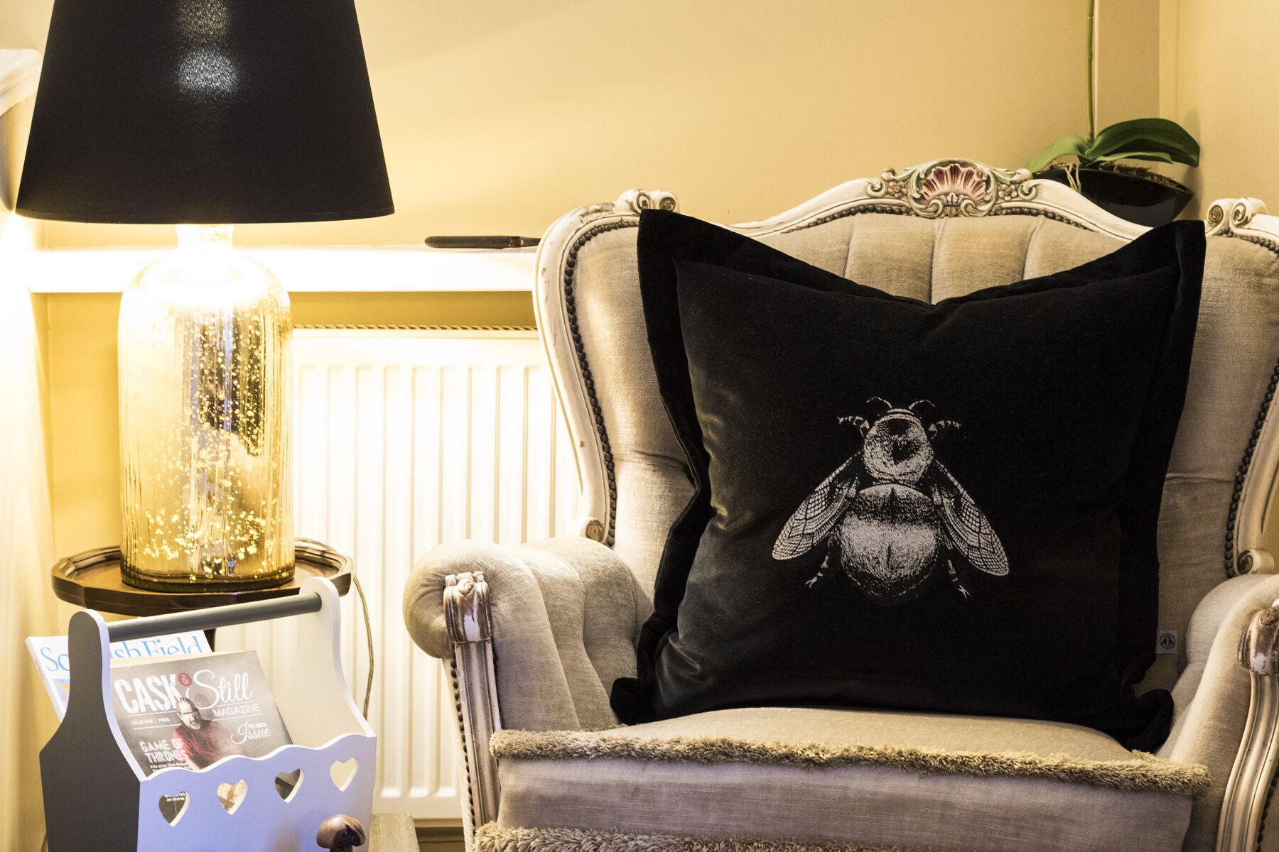 Ein Stuhl mit schwarzem Kissen mit Bär-Motiv, eine Lampe und ein Magazinregal mit einer Zeitschrift mit dem Titel "Cask & Still Magazine".